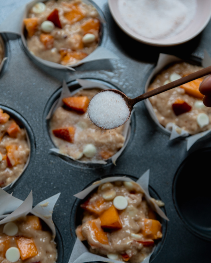 An overhead shot shows a spoon sprinkling some spiced sugar on top of muffin batter portions in a muffin tin.