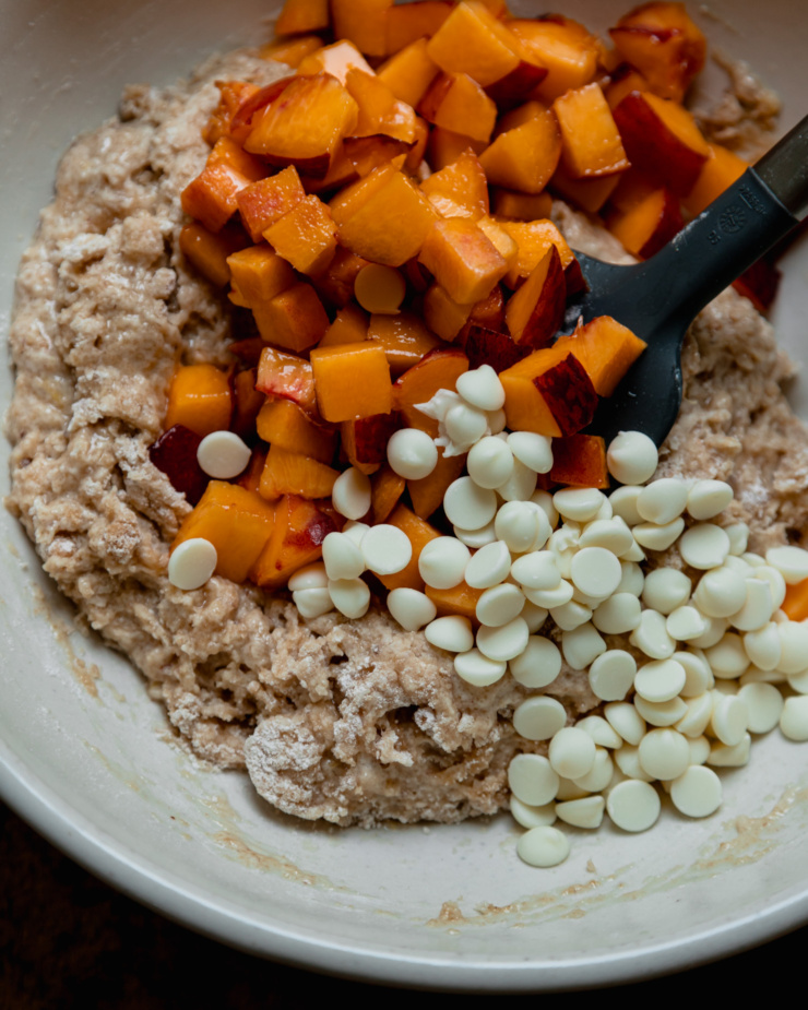 An overhead shot shows a bowl of mixed vegan muffin batter with chopped peaches and vegan white chocolate chips on top.