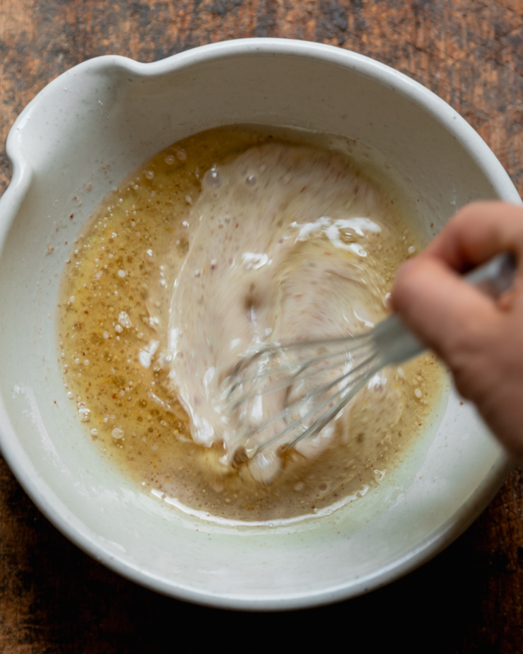 An overhead shot shows a hand using a whisk to stir up non-dairy milk, oil, lemon juice, ground flax, lemon zest, apple sauce, and vanilla extract together.