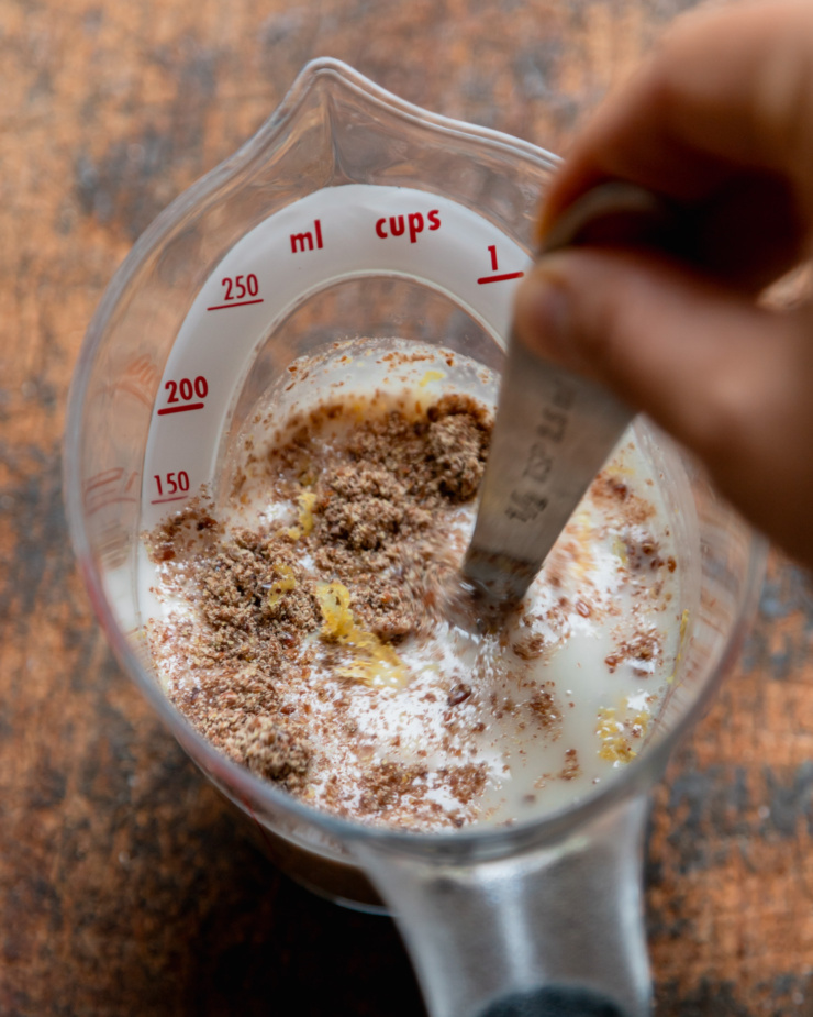 An overhead shot shows a hand using a measuring spoon to stir together soy milk, lemon zest, lemon juice, and ground flax seed together in a measuring cup.