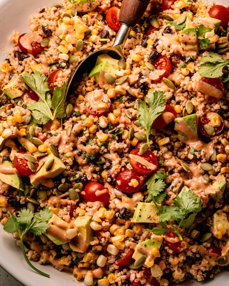 An overhead shot shows a charred corn quinoa salad in a wide bowl with a serving spoon sticking out. The salad features halved cherry tomatoes, black beans, avocado, cilantro, and a barbecue tahini ranch dressing.