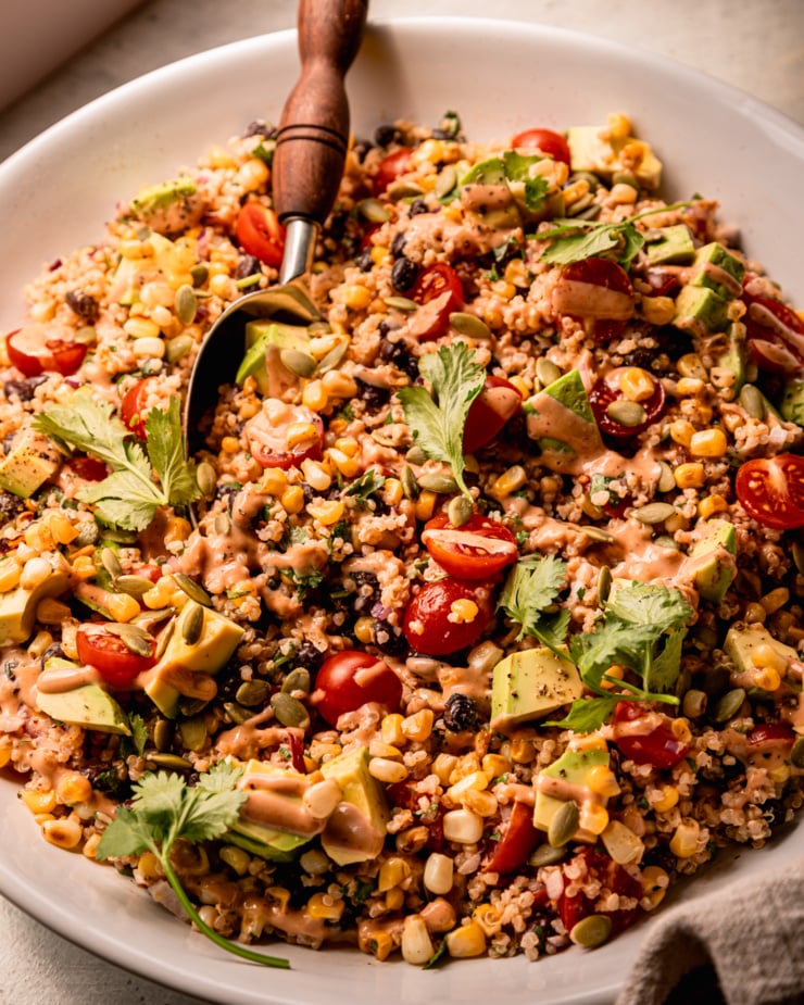 A 3/4 angle shot shows a charred corn quinoa salad in a wide bowl with a serving spoon sticking out. The salad features halved cherry tomatoes, avocado, cilantro, and a barbecue tahini ranch dressing.