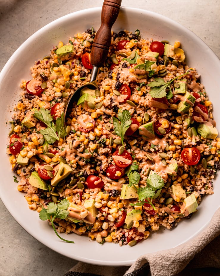 An up close overhead shot shows a charred corn quinoa salad in a wide bowl with a serving spoon sticking out. The salad features halved cherry tomatoes, avocado, cilantro, and a barbecue tahini ranch dressing.