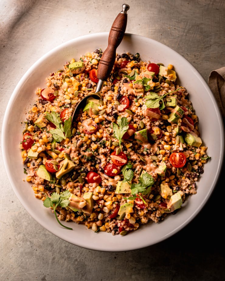 An overhead shot shows a charred corn quinoa salad in a wide bowl with a serving spoon sticking out. The salad features halved cherry tomatoes, avocado, cilantro, and a barbecue tahini ranch dressing.