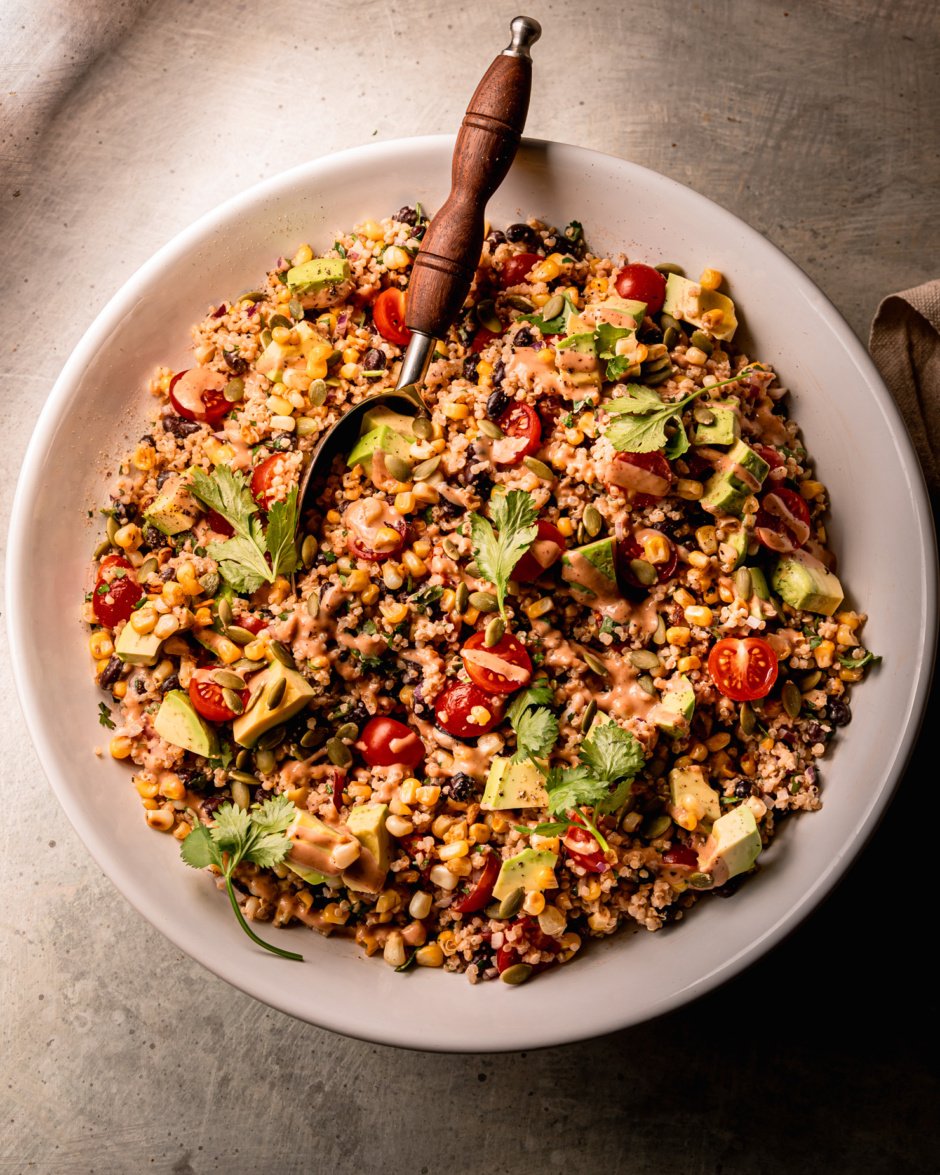 An overhead shot shows a charred corn quinoa salad in a wide bowl with a serving spoon sticking out. The salad features halved cherry tomatoes, avocado, cilantro, and a barbecue tahini ranch dressing.
