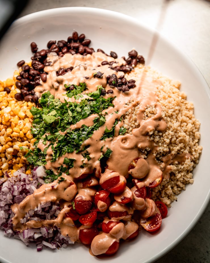 An overhead shot shows a charred corn quinoa salad being drizzled with barbecue tahini ranch dressing.
