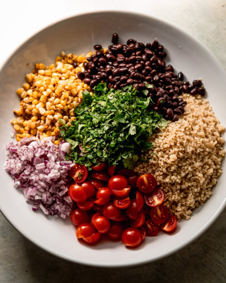 An overhead shot shows a wide bowl with segments of: black beans, cooked quinoa, halved cherry tomatoes, diced red onion, charred corn, and chopped cilantro.