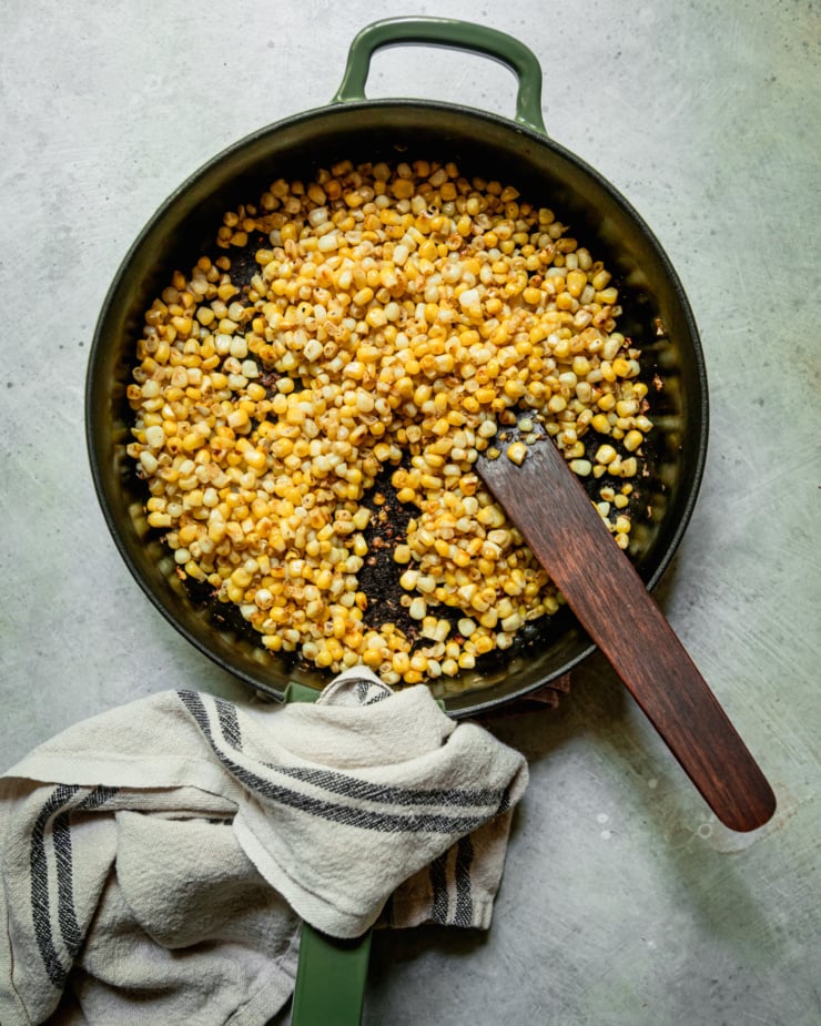 An overhead shot shows a cast iron skillet with cooked corn inside. A wooden stirring utensil is sticking out of the skillet.