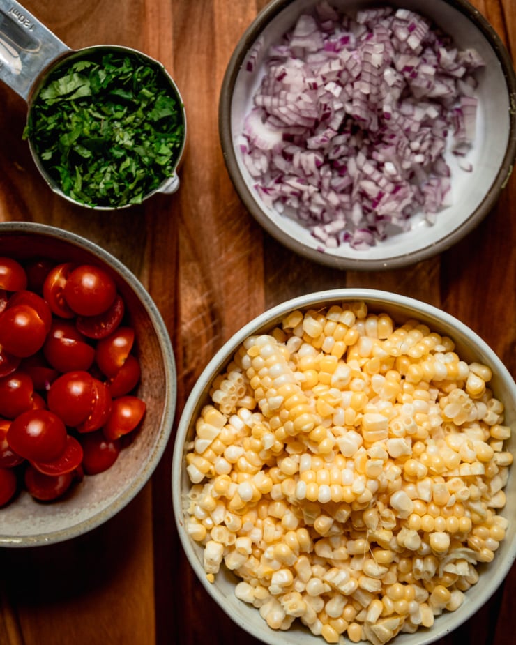 An overhead shot shows bowls of diced red onion, corn kernels, halved cherry tomatoes, and chopped cilantro.