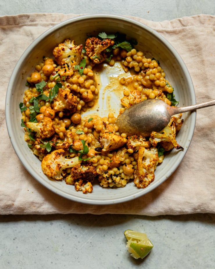 An overhead shot shows an individual serving of coconut pearl couscous with roasted cauliflower and chopped cilantro on top. The portion is partially eaten. Chickpeas are also seen in the saucy mixture. A spoon is sticking out of the side of the shallow bowl and a squeezed out lime wedge is nearby.