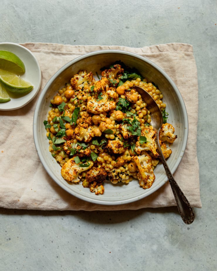 An overhead shot shows an individual serving of coconut pearl couscous with roasted cauliflower and chopped cilantro on top. Chickpeas are also seen in the saucy mixture. A spoon is sticking out of the shallow bowl.