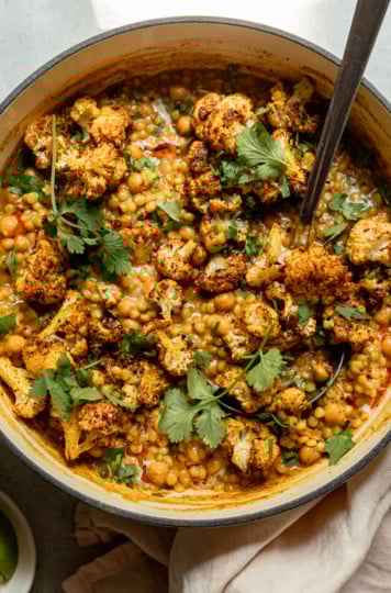 An overhead shot shows a braiser pot filled with spiced coconut pearl couscous, chickpeas, and roasted cauliflower florets. The dish has a stew-like texture and is garnished with cilantro.