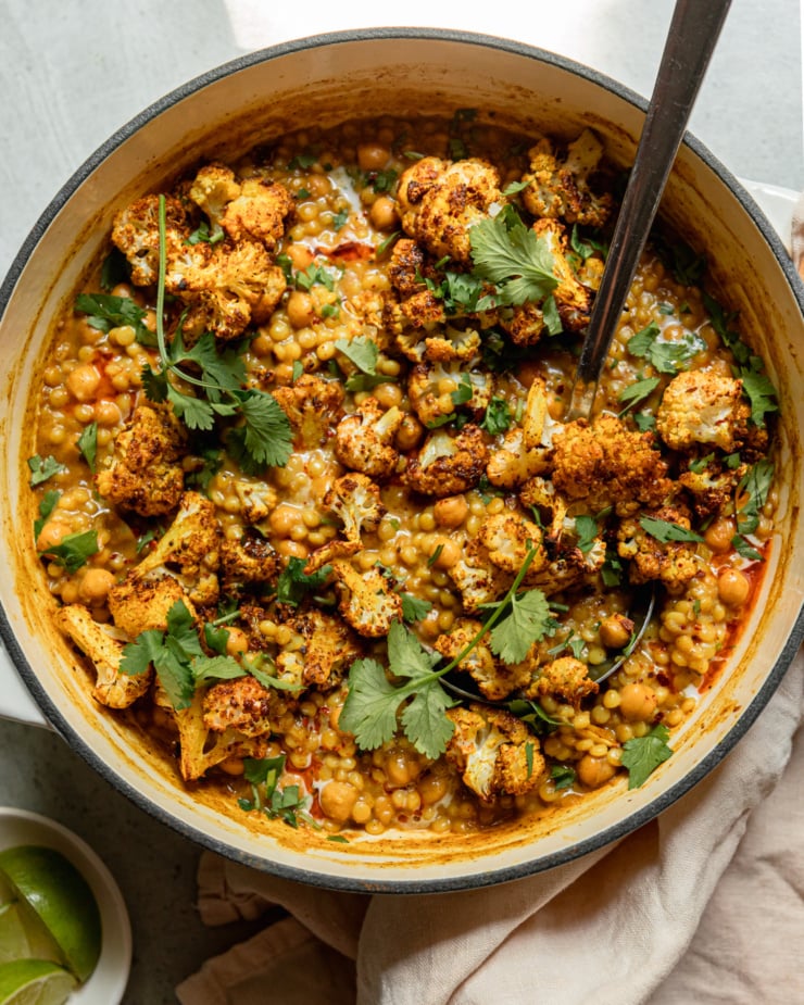 An overhead shot shows a braiser pot filled with spiced coconut pearl couscous, chickpeas, and roasted cauliflower florets. The dish has a stew-like texture and is garnished with cilantro.