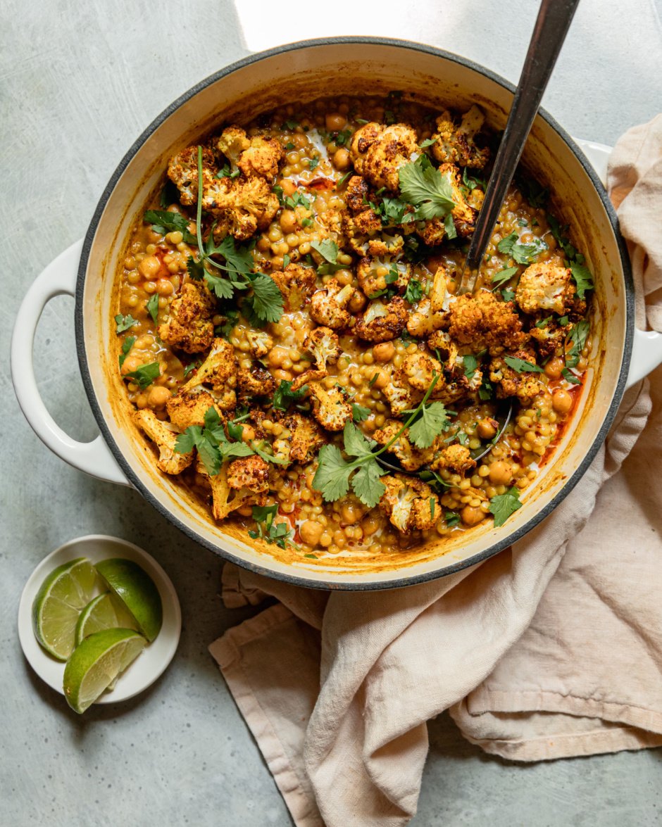 An overhead shot shows a braiser pot filled with spiced coconut pearl couscous, chickpeas, and roasted cauliflower florets. The dish has a stew-like texture and is garnished with cilantro. A small dish with lime wedges is nearby.