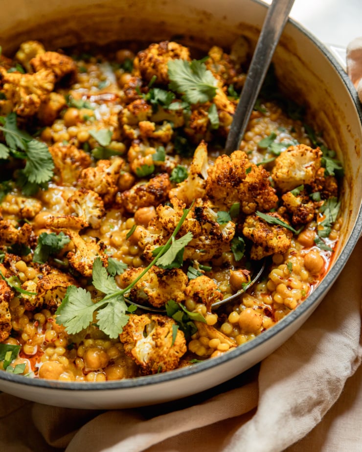 A 3/4 angle, up close shot shows a braiser pot with spiced pearl couscous, chickpeas, and roasted cauliflower. The dish is saucy with coconut milk and topped with chopped cilantro.