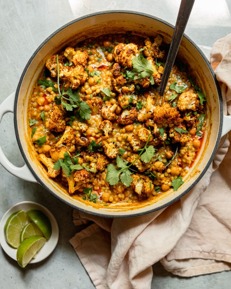 An overhead shot shows a braiser pot filled with spiced coconut pearl couscous, chickpeas, and roasted cauliflower florets. The dish has a stew-like texture and is garnished with cilantro. A small dish with lime wedges is nearby.