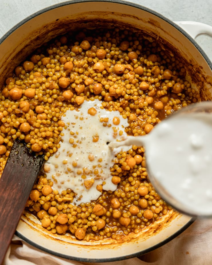 An overhead shot shows coconut milk being poured into a pot with cooked pearl couscous and chickpeas.
