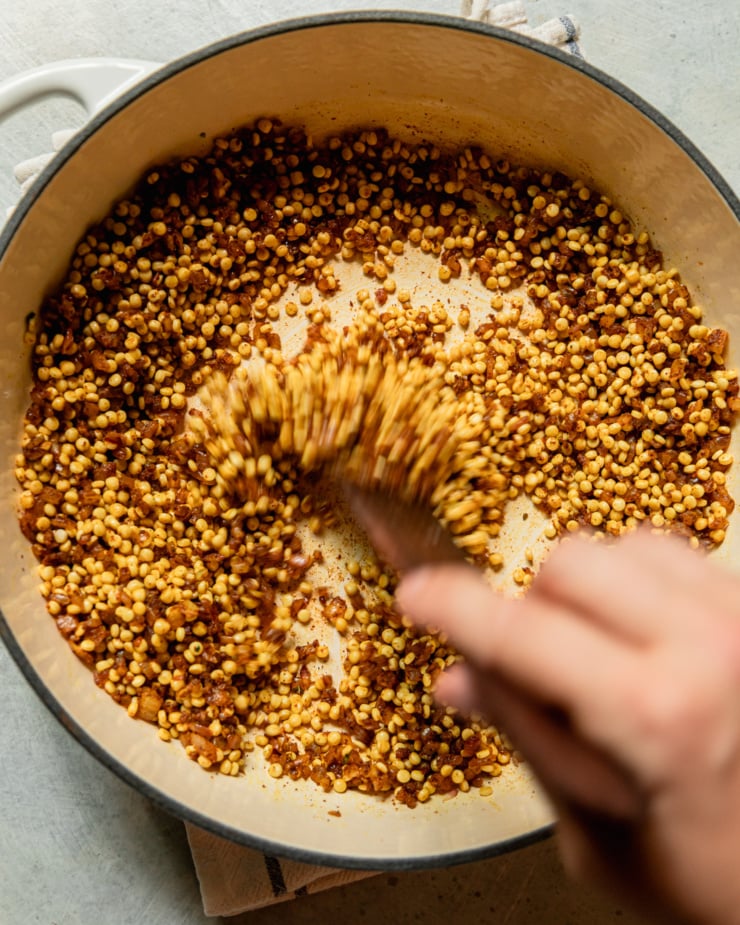 An overhead shot shows a hand using a wooden utensil to stir pearl couscous in a mixture of sautรฉed onions, minced garlic and spices.