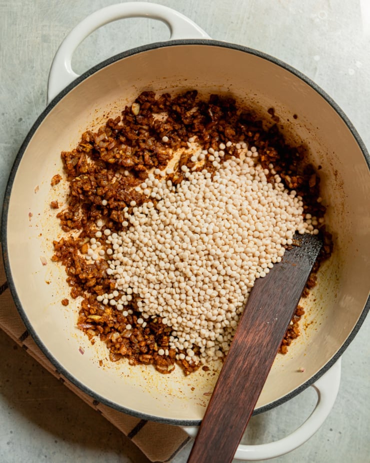 An overhead shot shows a braiser skillet filled with sautรฉed spiced onion and pearl couscous. A wooden stirring spatula is sticking out of the skillet.