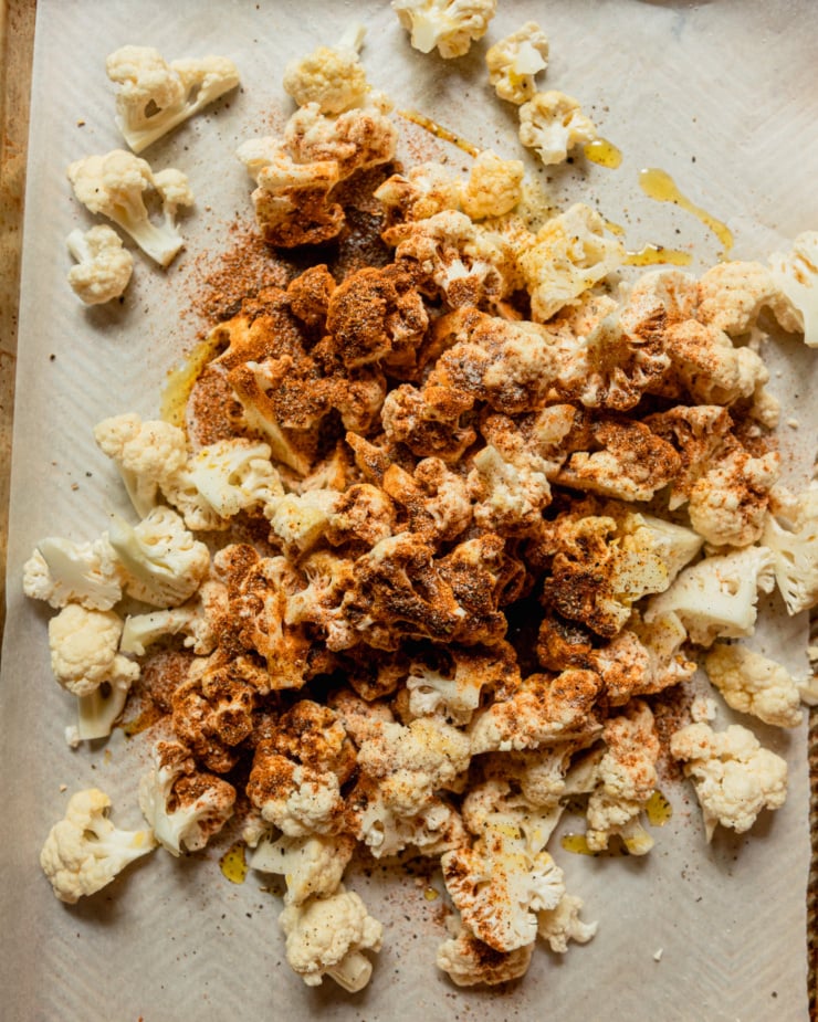 An overhead shot shows a pile of cauliflower florets on a baking sheet, drizzled with oil and dusted with spices.