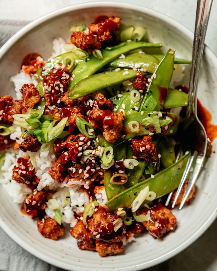 An overhead shot shows a bowl of crispy air fryer tempeh, cooked snow peas, and white rice. The food is drizzled with a gochujang glaze and garnished with sliced green onions and sesame seeds.