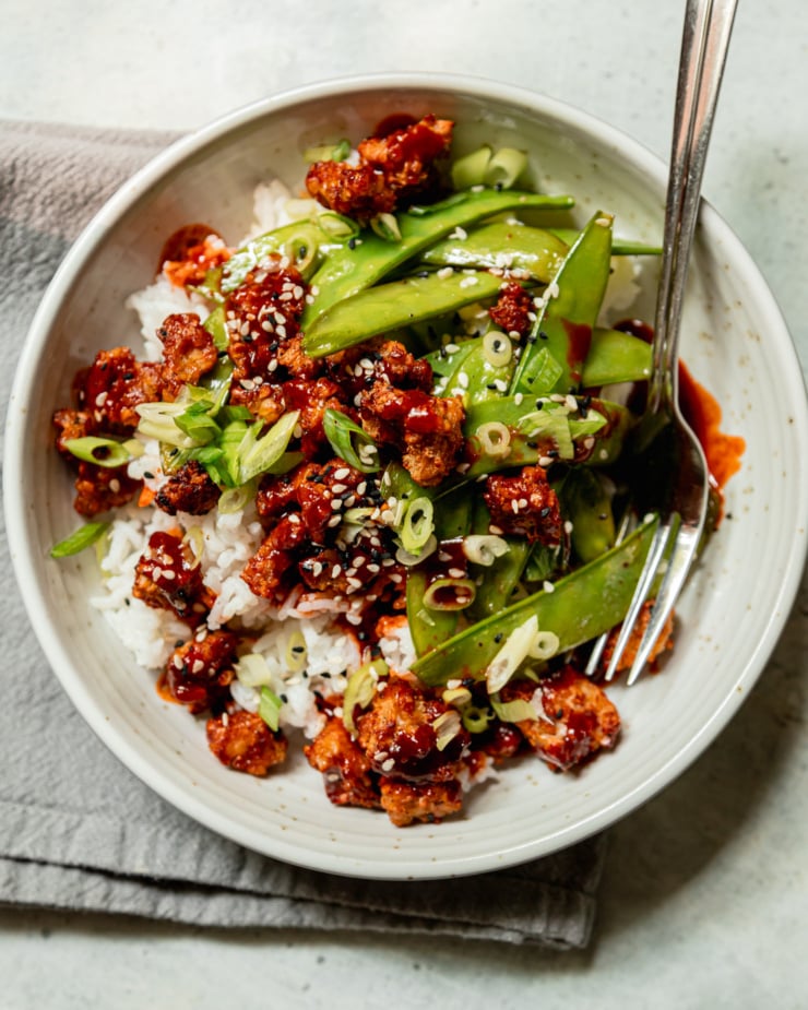 An overhead shot shows a bowl of crispy air fryer tempeh, cooked snow peas, and white rice. The food is drizzled with a gochujang glaze and garnished with sliced green onions and sesame seeds.