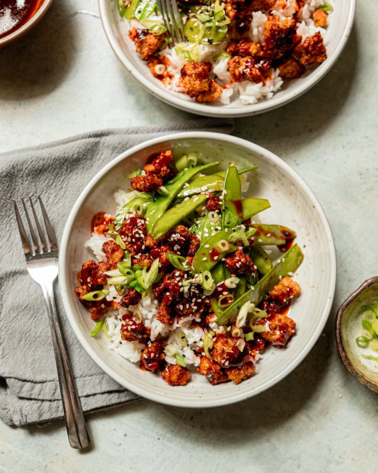 An overhead shot shows a bowl of crispy air fryer tempeh, cooked snow peas, and white rice. The food is drizzled with a gochujang glaze and garnished with sliced green onions and sesame seeds.