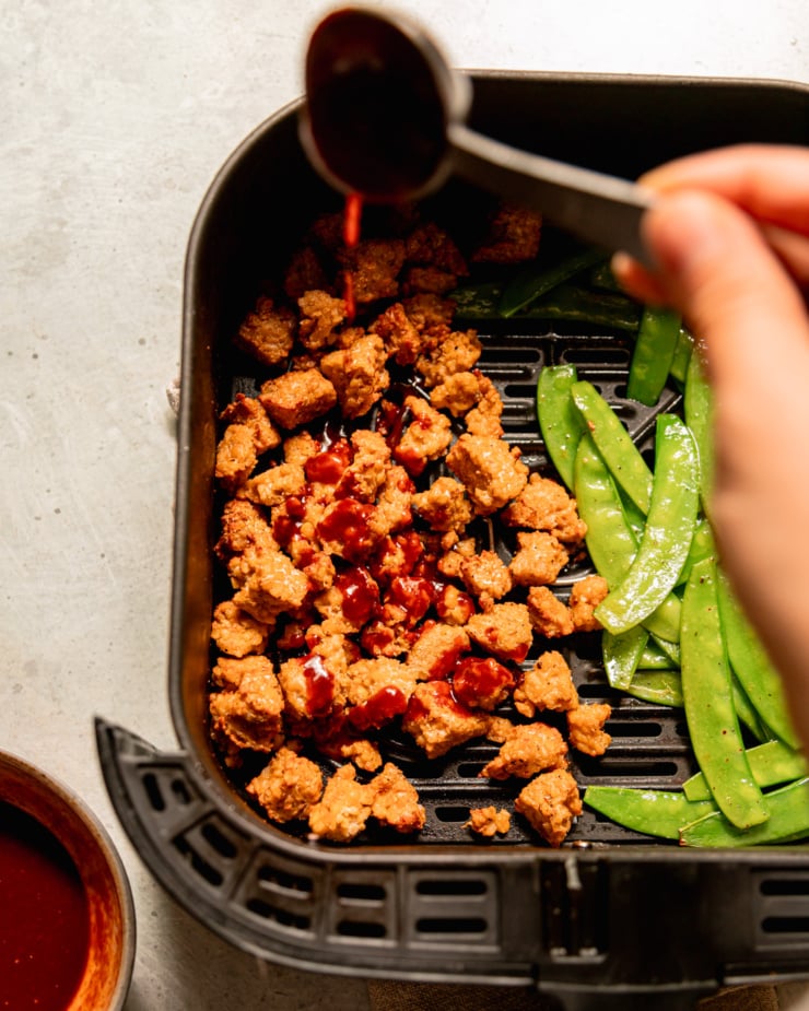 An overhead shot shows a hand using a spoon to drizzle gochujang glaze over some tempeh bits in an air fryer basket. Snow peas are seen alongside.