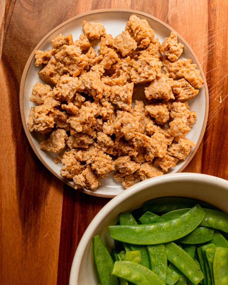 An overhead shot shows a plate filled with torn tempeh pieces and a bowl with trimmed raw snow peas.