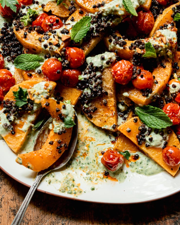 An overhead shot shows a platter with toasted butternut squash slices, burst cherry tomatoes, herbed creamy vegan yogurt sauce, roasted chewy lentils, whole herbs, and drizzles of chili oil. A serving spoon has taken a portion away, leaving chili oil swirls in the yogurt sauce.