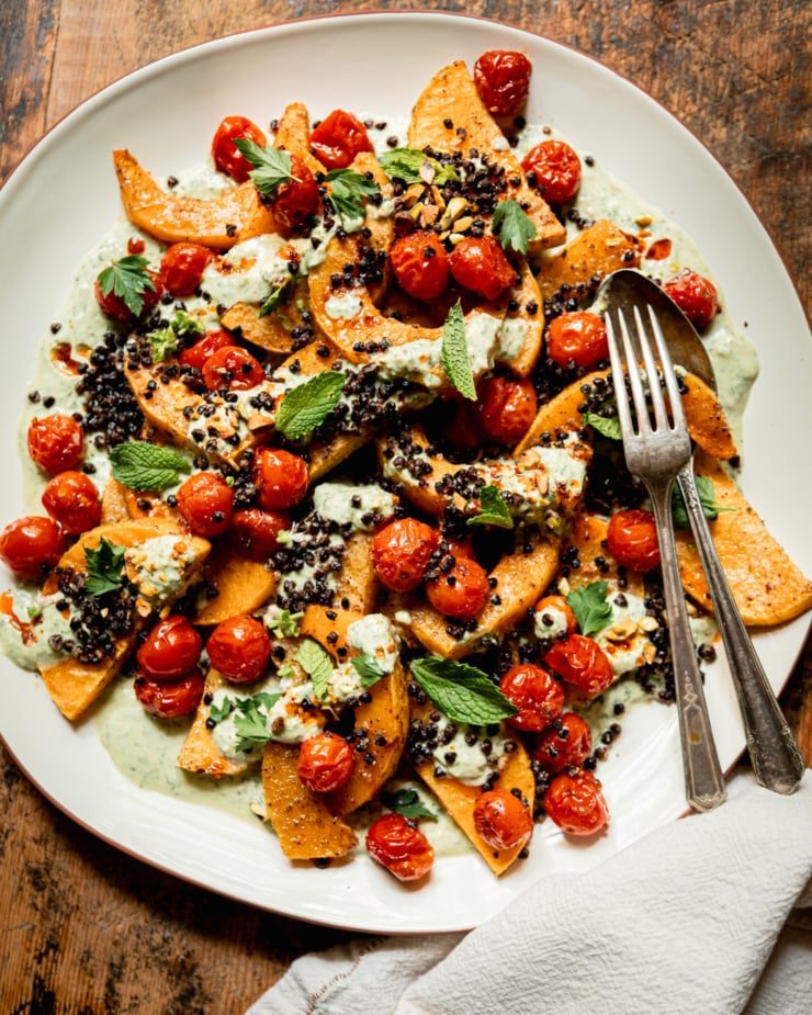 An overhead shot shows a platter with toasted butternut squash slices, burst cherry tomatoes, herbed creamy vegan yogurt sauce, roasted chewy lentils, whole herbs, and drizzles of chili oil. Serving utensils are seen to the side.