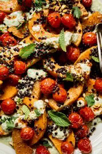 An up close, overhead shot shows a platter with toasted butternut squash slices, burst cherry tomatoes, herbed creamy vegan yogurt sauce, roasted chewy lentils, whole herbs, and drizzles of chili oil. Serving utensils are seen to the side.
