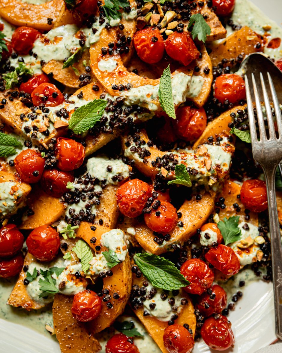 An up close, overhead shot shows a platter with toasted butternut squash slices, burst cherry tomatoes, herbed creamy vegan yogurt sauce, roasted chewy lentils, whole herbs, and drizzles of chili oil. Serving utensils are seen to the side.