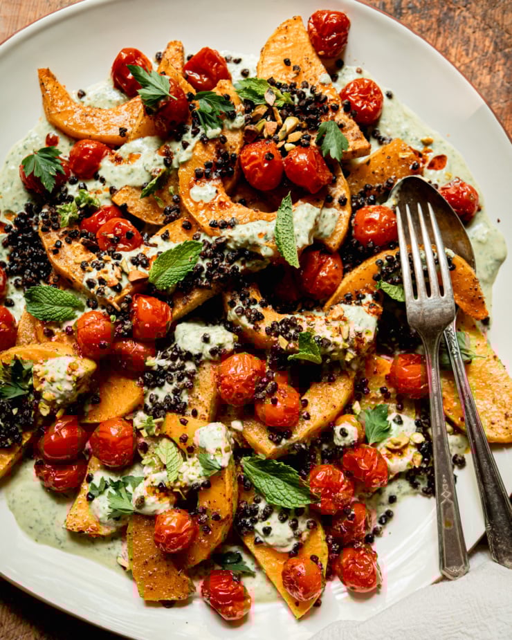 An overhead shot shows a platter with toasted butternut squash slices, burst cherry tomatoes, herbed creamy vegan yogurt sauce, roasted chewy lentils, whole herbs, and drizzles of chili oil. Serving utensils are seen to the side.