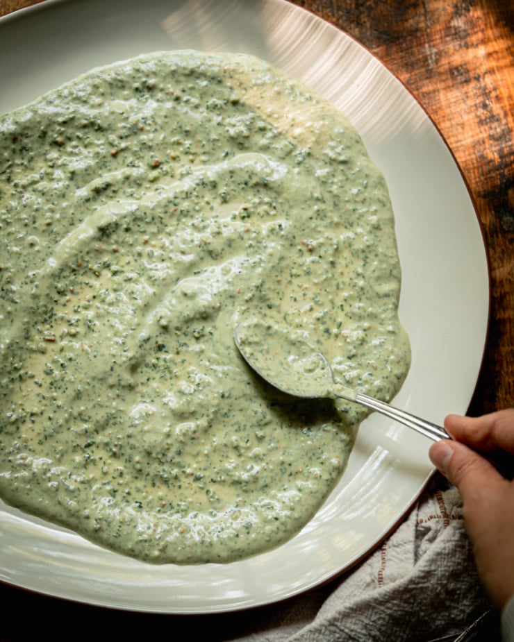 An overhead shot shows a hand using a spoon to spread a pale green vegan yogurt sauce out onto the base of a serving platter.