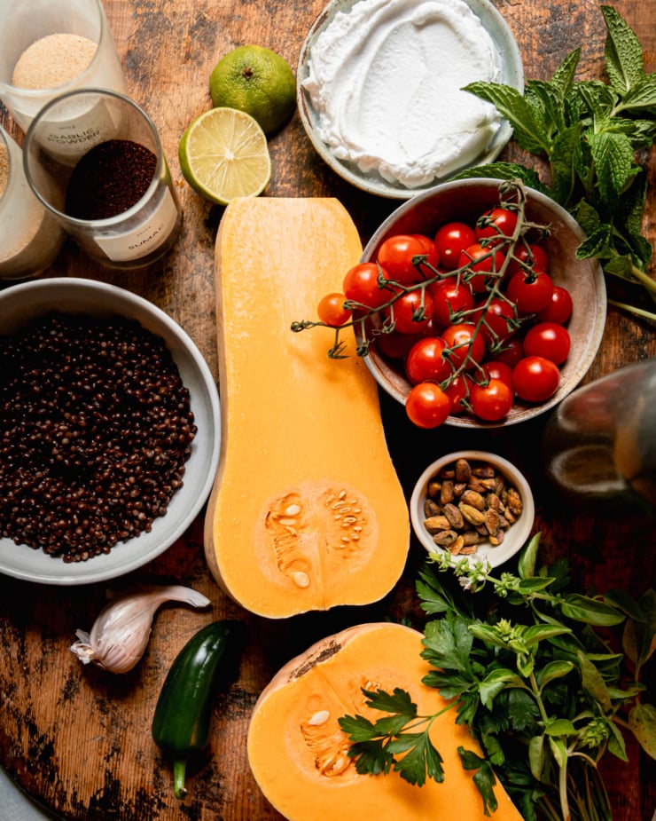 An overhead shot shows ingredients for a vegan recipe: butternut squash, herbs, tomatoes, lentils, vegan yogurt, lime, spices, pistachios, jalapeño and garlic.