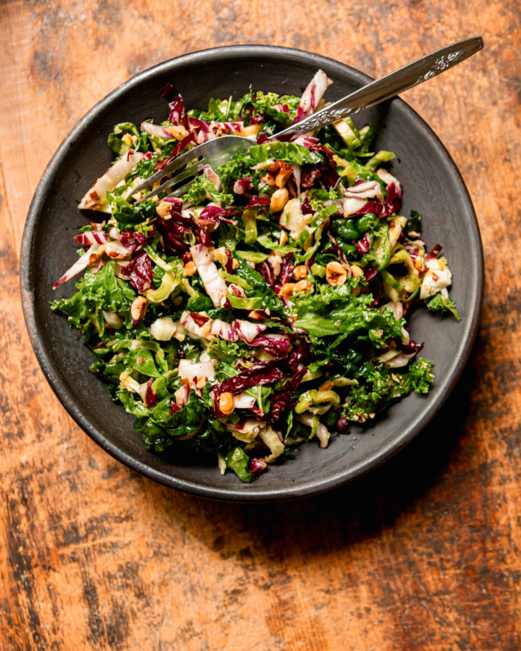 An overhead shot shows an individual serving of a shredded fall greens salad topped with chopped hazelnuts. A fork is sticking out of the bowl.