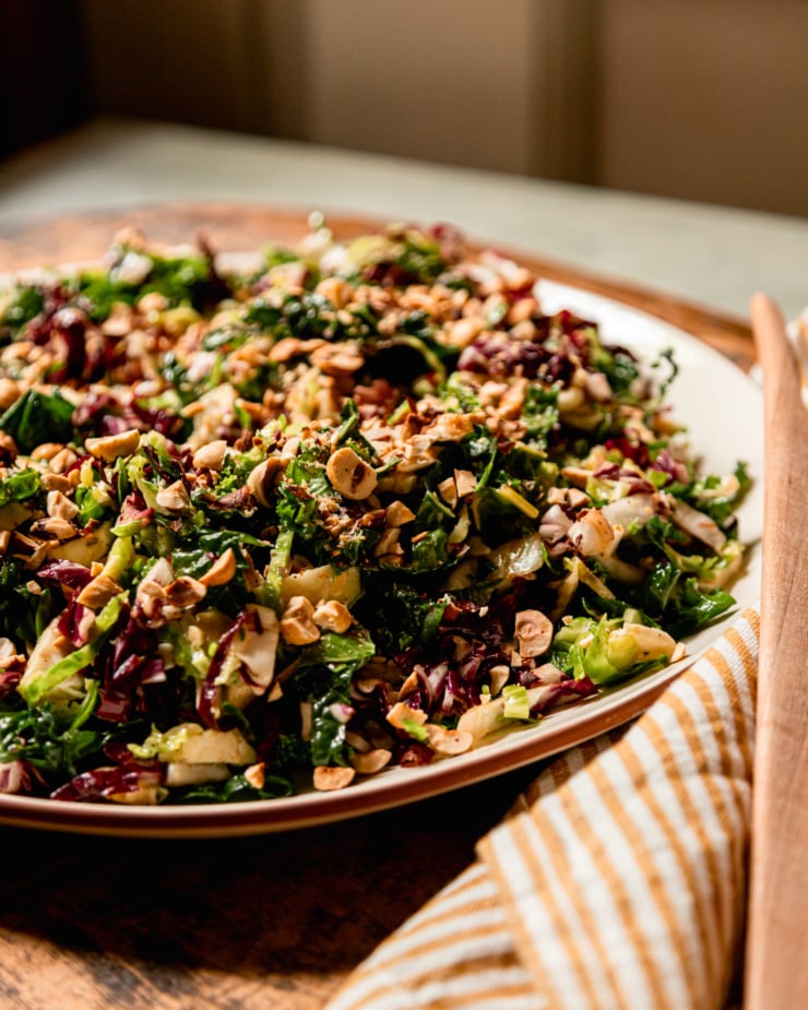 A head-on shot shows a platter filled with a shredded fall greens salad. The dressed greens are topped with chopped roasted hazelnuts.
