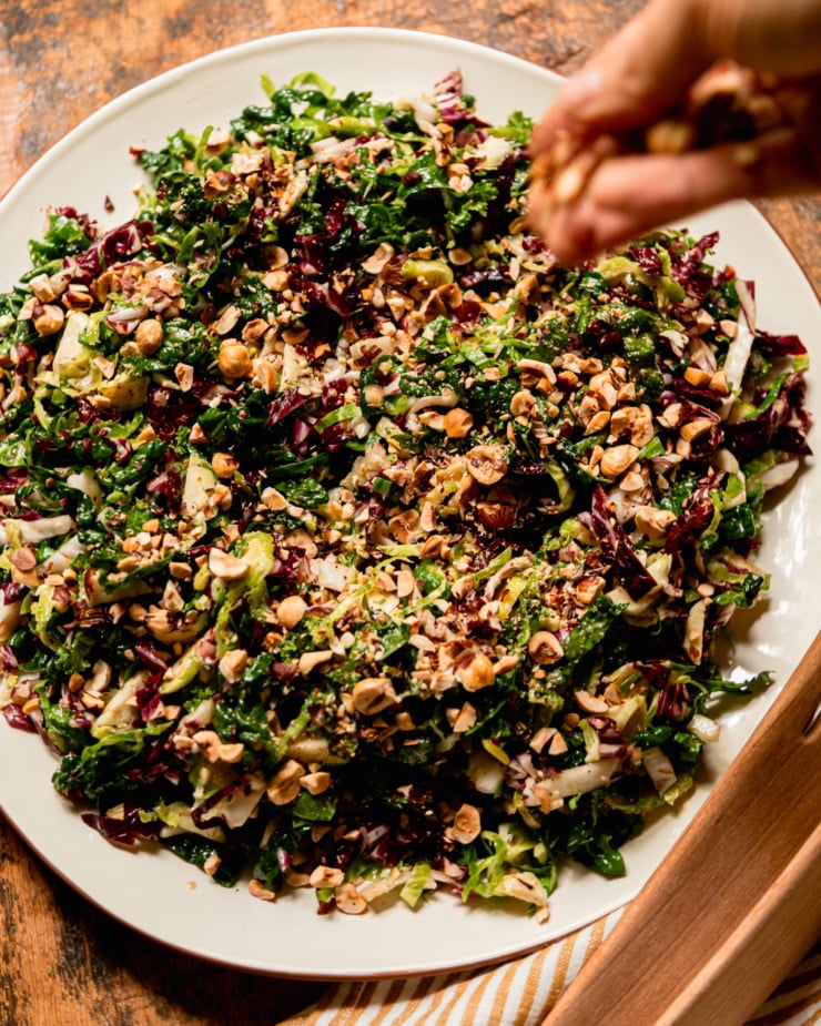 An overhead shot shows a hand scattering chopped hazelnuts over a shredded fall greens salad with sherry thyme vinaigrette.