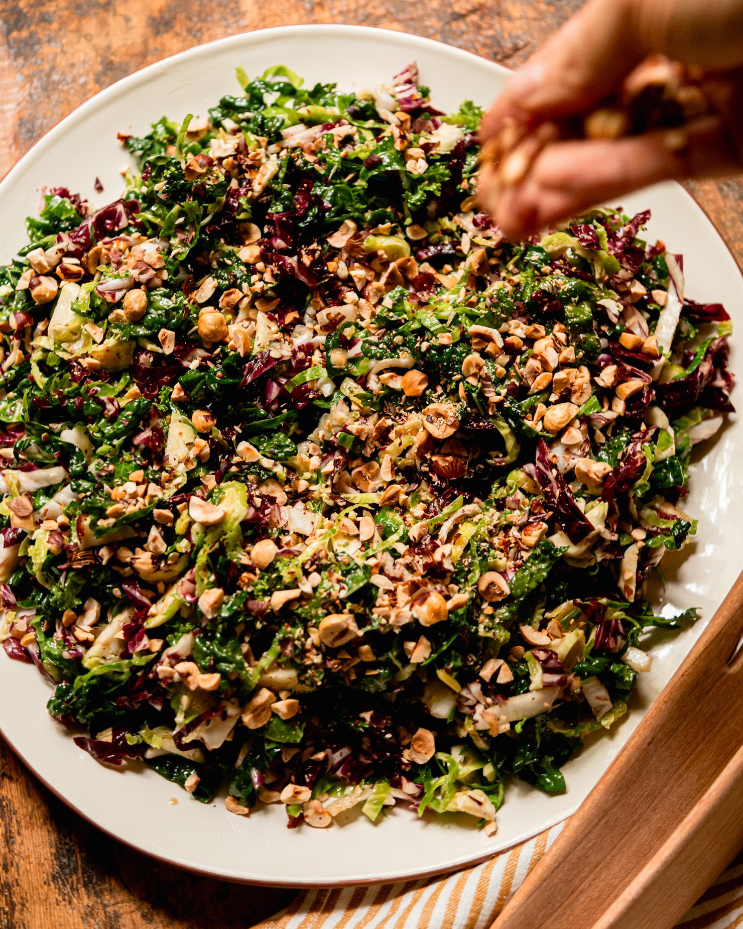 An overhead shot shows a hand scattering chopped hazelnuts over a shredded fall greens salad with sherry thyme vinaigrette.