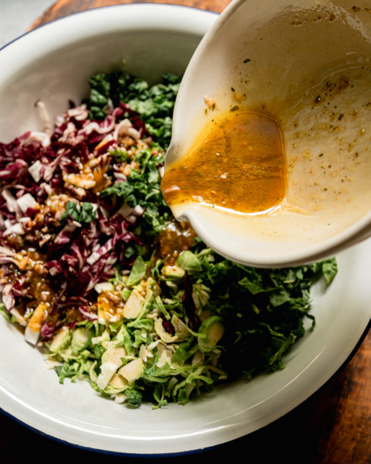 An overhead shot shows a vinaigrette being poured on top of shredded radicchio, kale, and brussels sprouts.