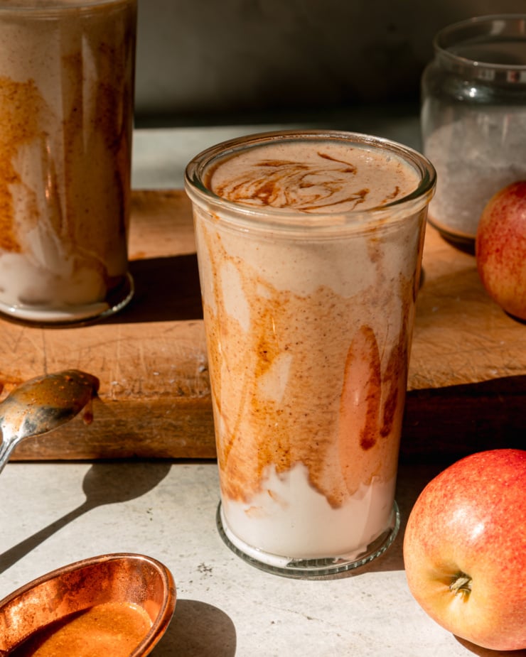 A head-on shot shows a glass of salted caramel apple smoothie. The glass has been drizzled with almond butter "caramel" sauce and has a swoop of vegan yogurt at the bottom. Apples are seen nearby; all on a wooden board.
