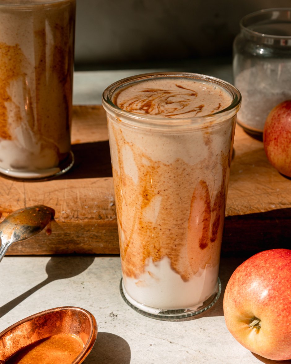 A head-on shot shows a glass of salted caramel apple smoothie. The glass has been drizzled with almond butter "caramel" sauce and has a swoop of vegan yogurt at the bottom. Apples are seen nearby; all on a wooden board.