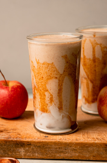 A head-on shot shows two glasses of salted caramel apple smoothie. The glasses have been drizzled with almond butter "caramel" sauce and have swoops of vegan yogurt at the bottom. Apples are seen nearby; all on a wooden board.