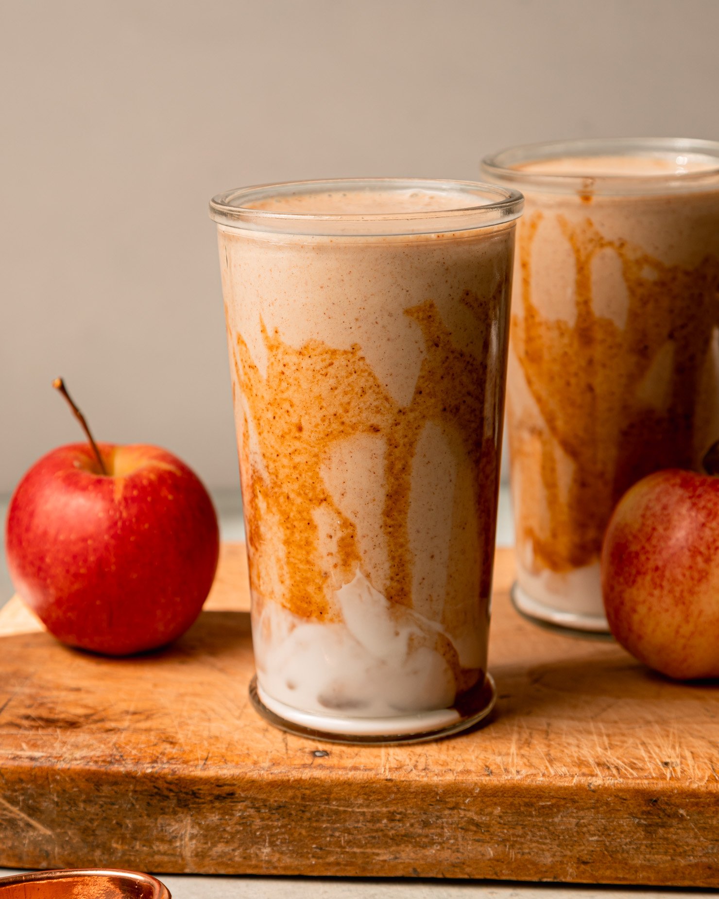 A head-on shot shows two glasses of salted caramel apple smoothie. The glasses have been drizzled with almond butter "caramel" sauce and have swoops of vegan yogurt at the bottom. Apples are seen nearby; all on a wooden board.