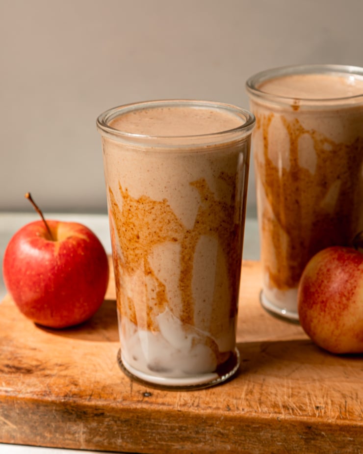 A head-on shot shows two glasses of salted caramel apple smoothie. The glasses have been drizzled with almond butter "caramel" sauce and have swoops of vegan yogurt at the bottom. Apples are seen nearby; all on a wooden board.