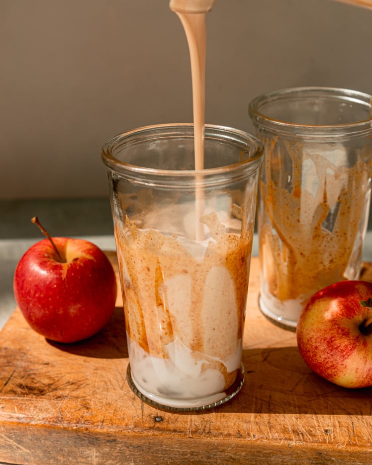 A head-on shot shows a salted caramel apple smoothie being poured into a glass that's been drizzled with almond butter "caramel" sauce. Apples are seen nearby.