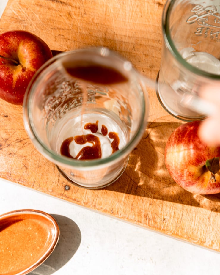 An overhead, slightly blurry shot shows a spoon drizzling an almond butter "caramel" sauce into the base of a drinking glass.