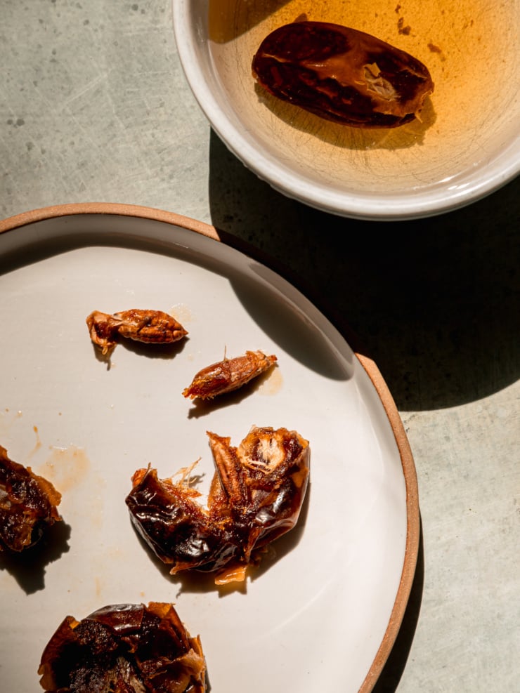 An overhead shot shows soaked dates that have been pitted on a plate. A date is still soaking in water to the side.