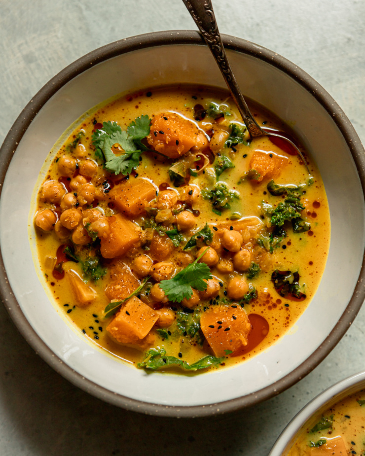 An overhead shot shows an individual serving of a ginger squash coconut stew with chickpeas and kale. A spoon is sticking out of the bowl.